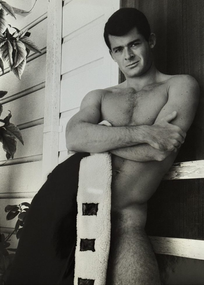 A black and white photograph of a nude man, covering his lower body with a jacket.