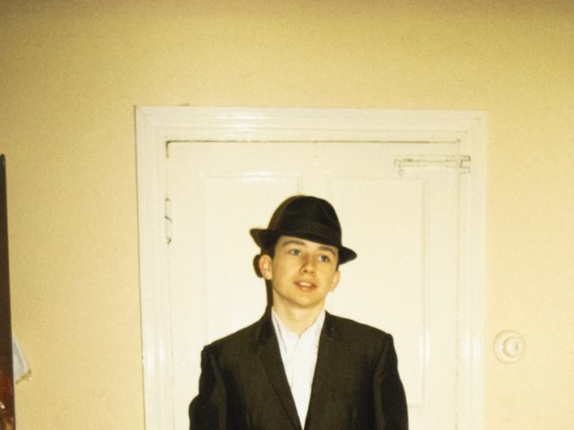 A photograph of a young boy in a dark suit and fedora standing in front of a doorway in a yellow room with a black and white dog playing by his feet.