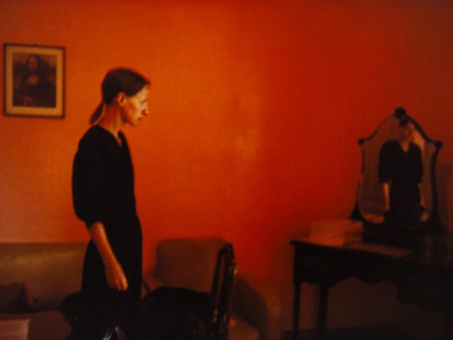 A woman stands in a room with red walls and looks over at a mirrored vanity in the corner.