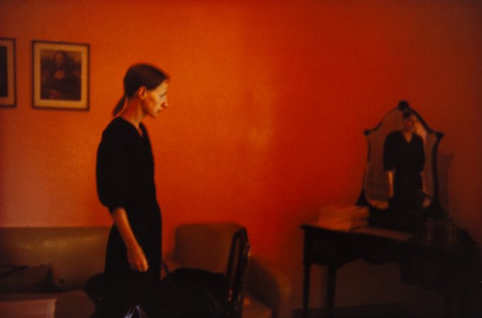 A woman stands in a room with red walls and looks over at a mirrored vanity in the corner.