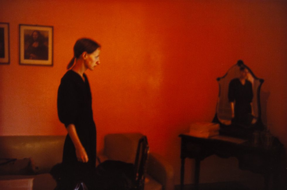 A woman stands in a room with red walls and looks over at a mirrored vanity in the corner.