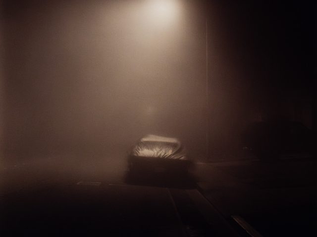 A moody image of a car at night in fog under a streetlight.