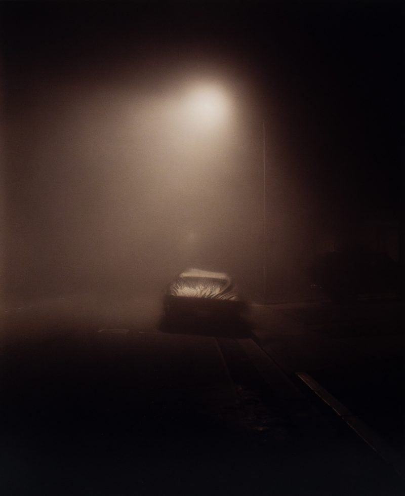 A moody image of a car at night in fog under a streetlight.