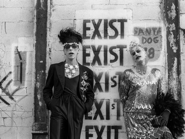 A black and white photograph of two people in flamboyant outfits standing in front of a wall with grafitti.