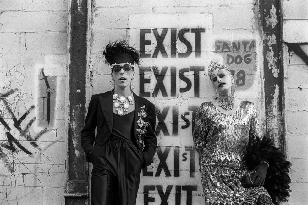 A black and white photograph of two people in flamboyant outfits standing in front of a wall with grafitti.