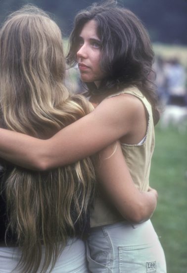 A color photograph of two women with long hair embracing in a green field.
