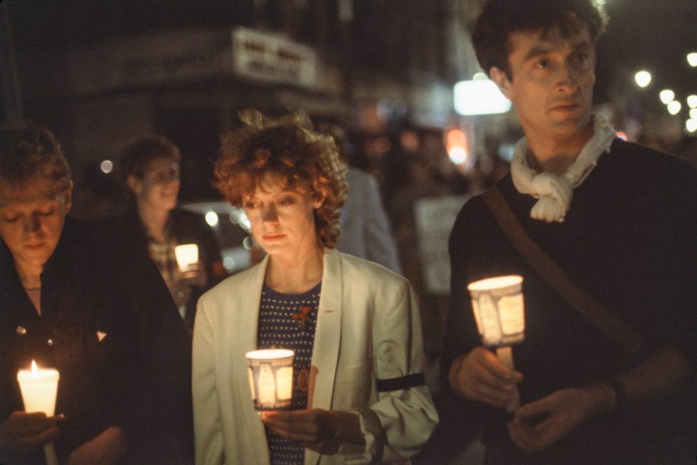 A color photograph of three individuals holding candles at a vigil/march for AIDS victims.