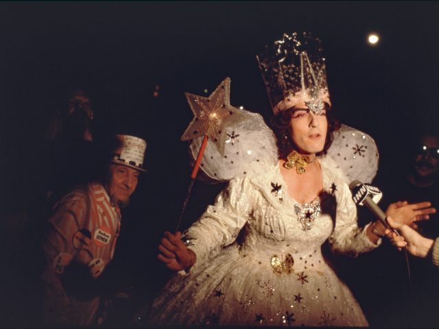 A color photograph of a woman in a fairy dress holding a magic wand being interviewed by a news crew.