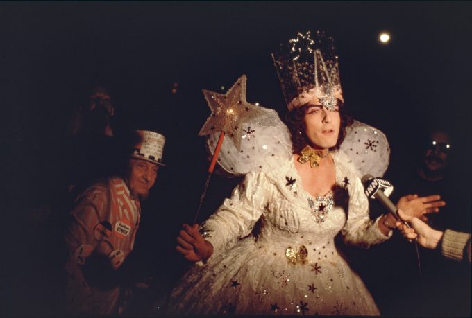A color photograph of a woman in a fairy dress holding a magic wand being interviewed by a news crew.