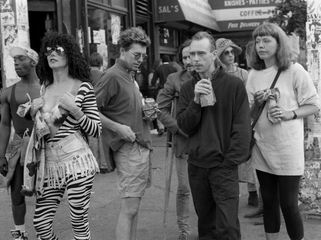 A black and white photograph of people standing around on a city sidewalk.