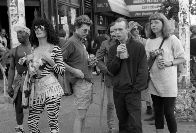 A black and white photograph of people standing around on a city sidewalk.