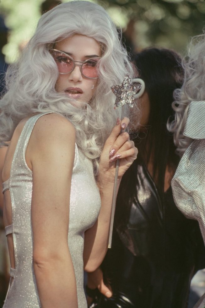 A color photograph of a woman dressed as a fairy with sunglasses and a wand.