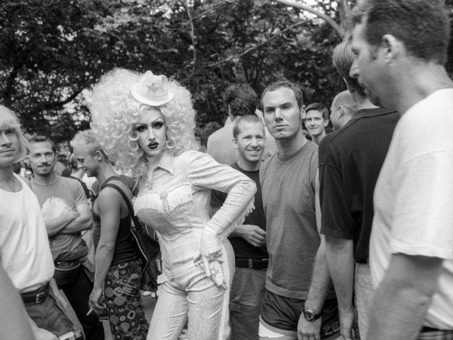 A black and white photograph of someone in a large blond wig and country western clothing in a crowd of gay men.