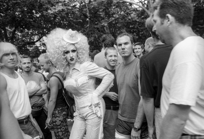 A black and white photograph of someone in a large blond wig and country western clothing in a crowd of gay men.