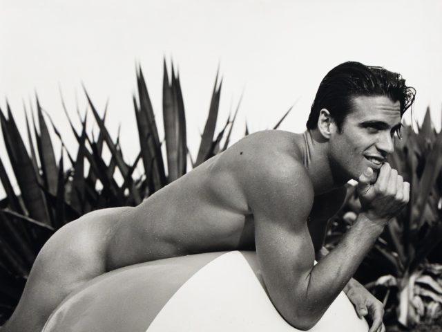 A black and white photograph of a nude man on a large inflatable beach ball.