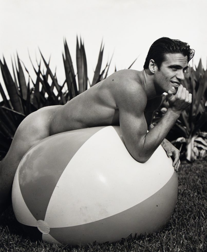 A black and white photograph of a nude man on a large inflatable beach ball.
