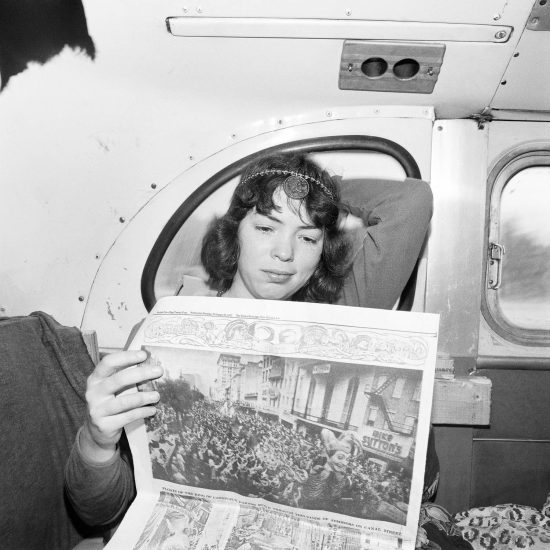 A black and white photograph of a young woman reading a newspaper on a bus.