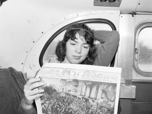 A black and white photograph of a young woman reading a newspaper on a bus.