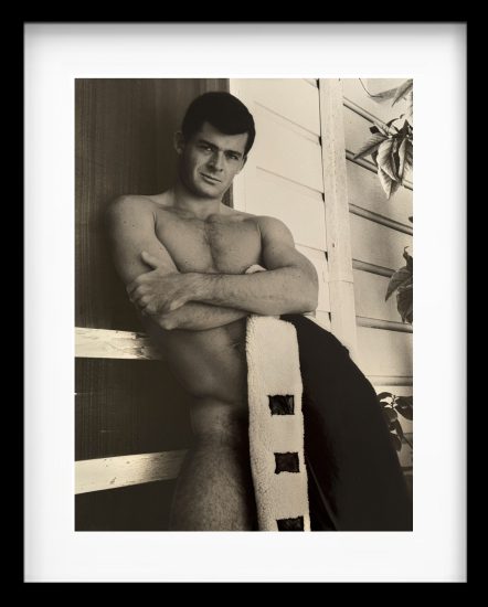 A black-and-white photograph of a handsome man in the nude with a jacket covering his intimate region.