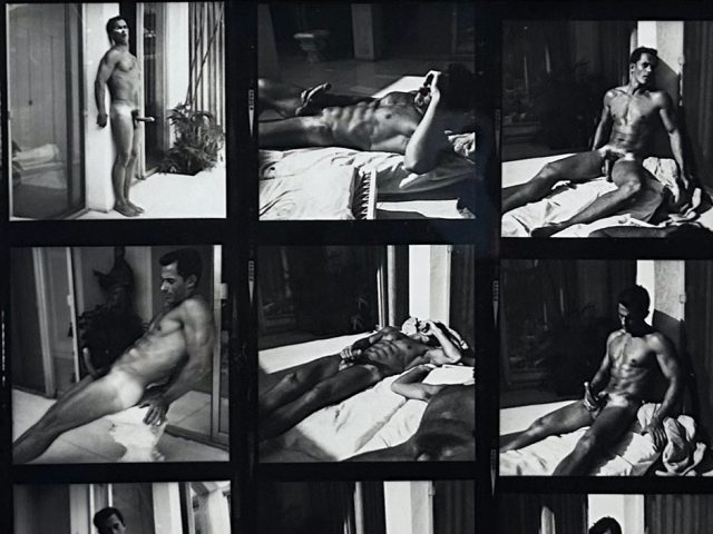 A black and white photograph of a nude man sitting outside in the sun.