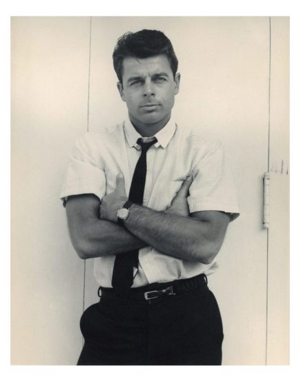 A black and white portrait of a man with his arms crossed in a short sleeve white shirt with a black tie.