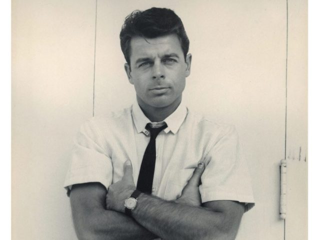 A black and white portrait of a man with his arms crossed in a short sleeve white shirt with a black tie.