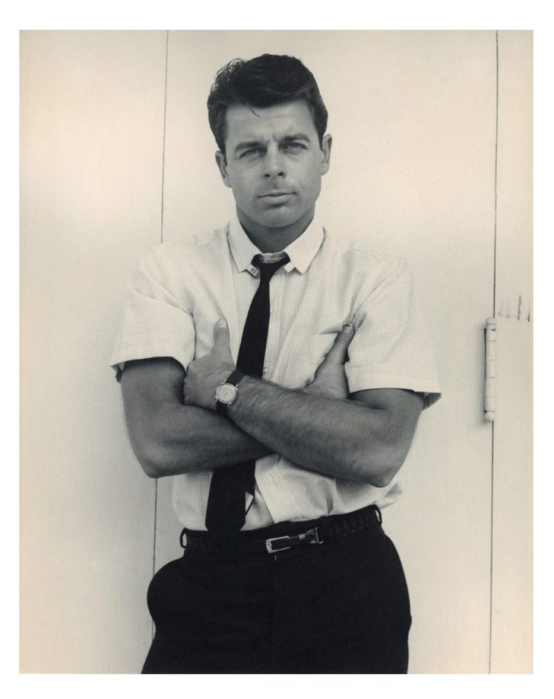 A black and white portrait of a man with his arms crossed in a short sleeve white shirt with a black tie.