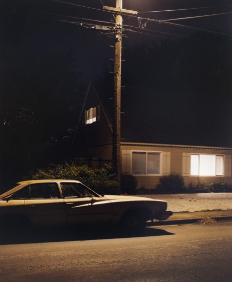 A color photograph of a car next to a house at night.