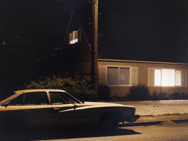 A color photograph of a car next to a house at night.