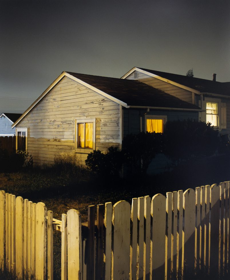 A photograph of a house with a white picket fence photographed at night with windows illuminated.