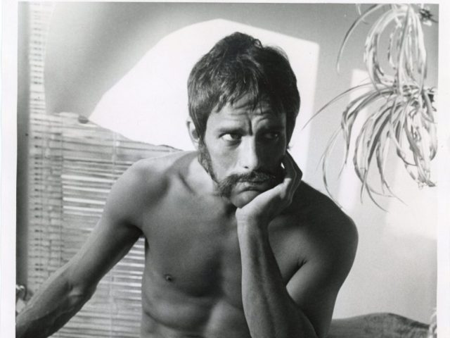 A black and white photograph of a nude man with his elbow resting in his knee.