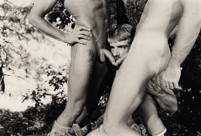 A black-and-white photograph of two nude men sticking their genitals in another man's face.