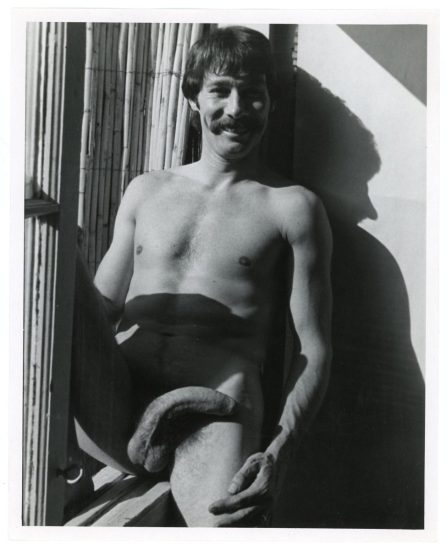 A black and white photograph of a nude man sitting by a window.