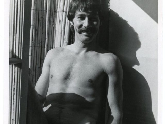 A black and white photograph of a nude man sitting by a window.