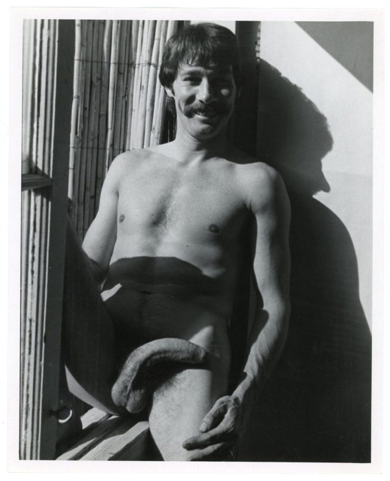 A black and white photograph of a nude man sitting by a window.