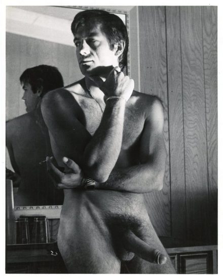 A black and white photograph of a nude man with an erection standing in a room with wood panelling.