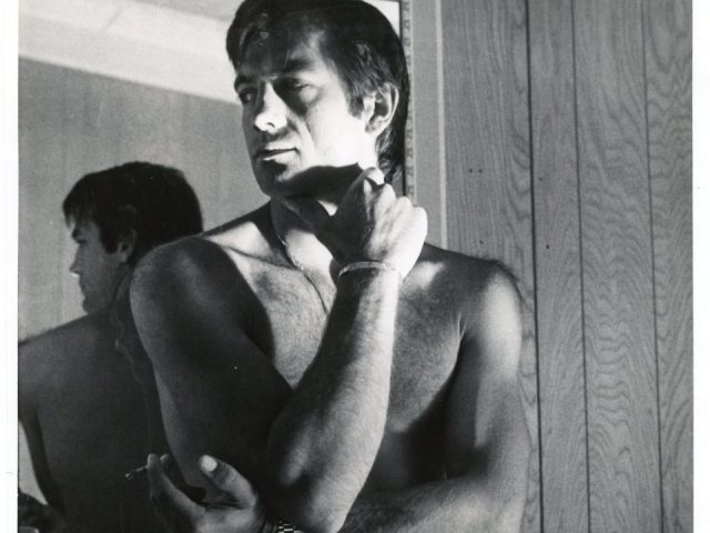 A black and white photograph of a nude man with an erection standing in a room with wood panelling.