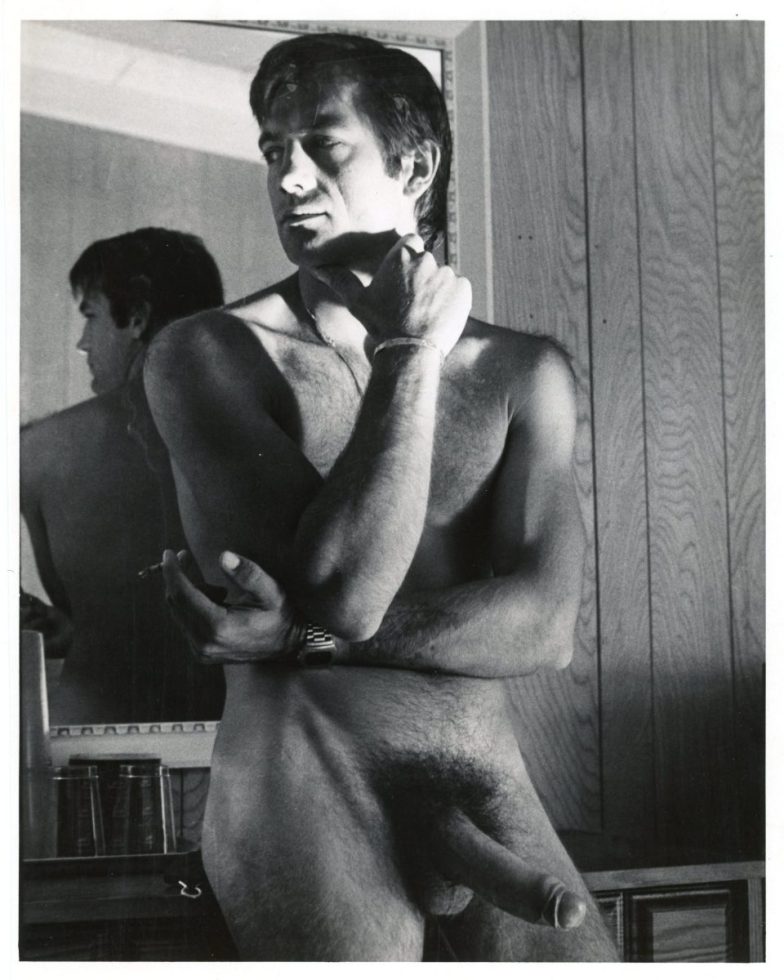 A black and white photograph of a nude man with an erection standing in a room with wood panelling.