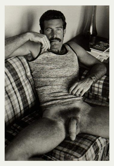 A black and white photograph of a nude man sitting on a plaid sofa.