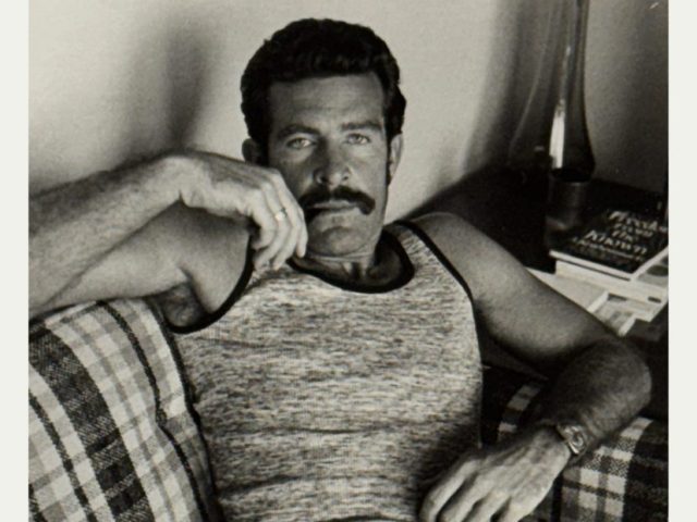 A black and white photograph of a nude man sitting on a plaid sofa.