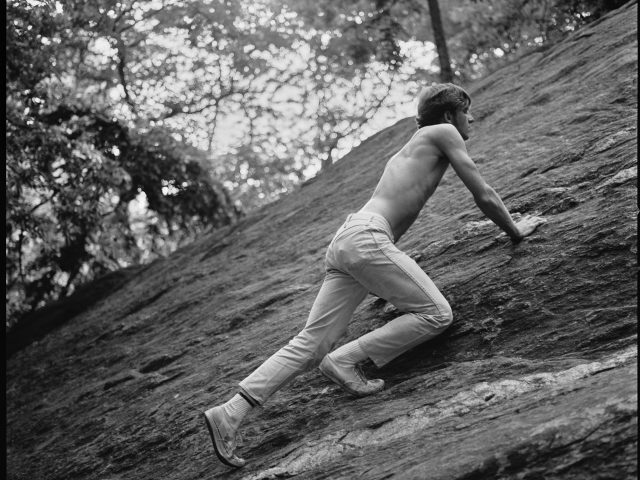 A black and white photograph of a shirtless man climbing a boulder.