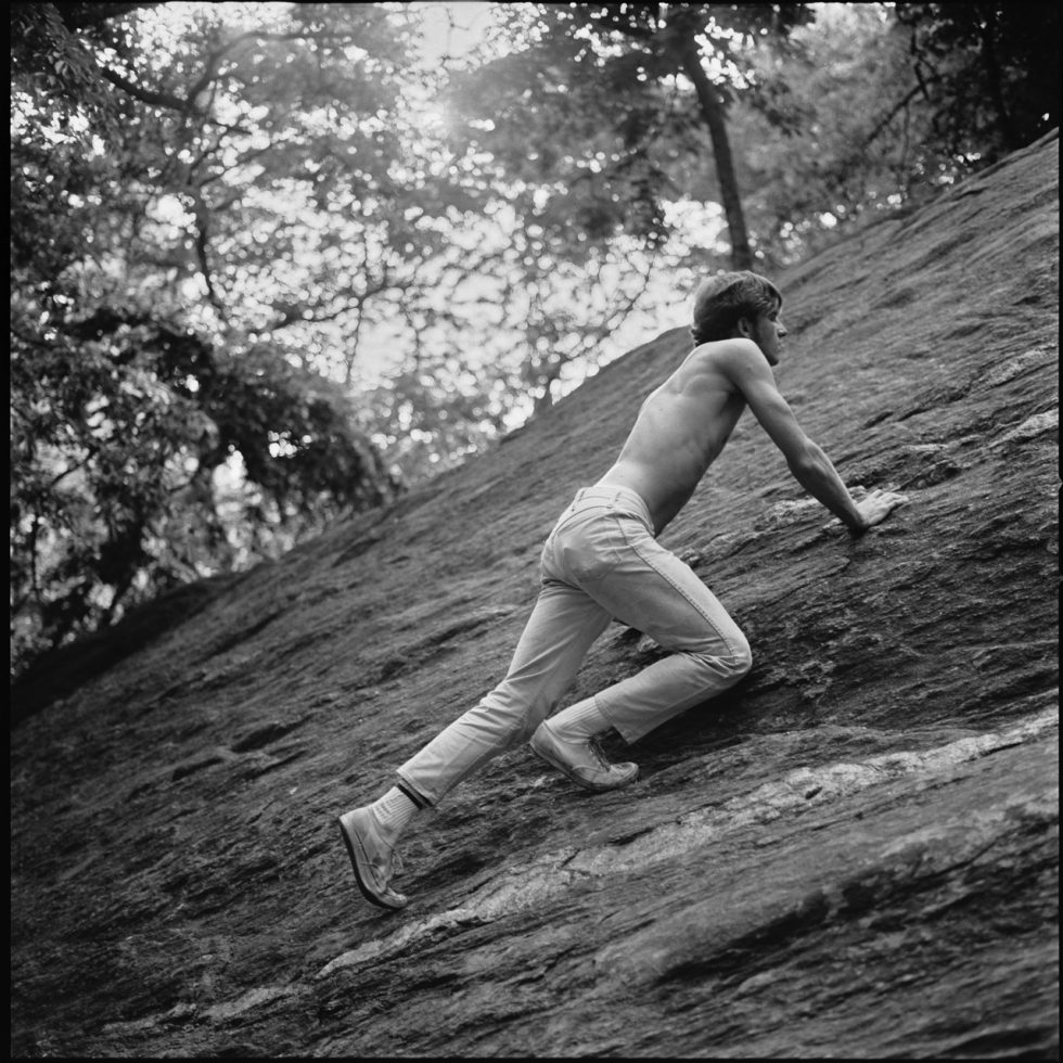 A black and white photograph of a shirtless man climbing a rock.