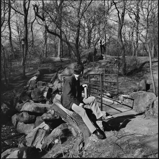 A black and white photograph of a young man sitting on a rock in a park holding a book.