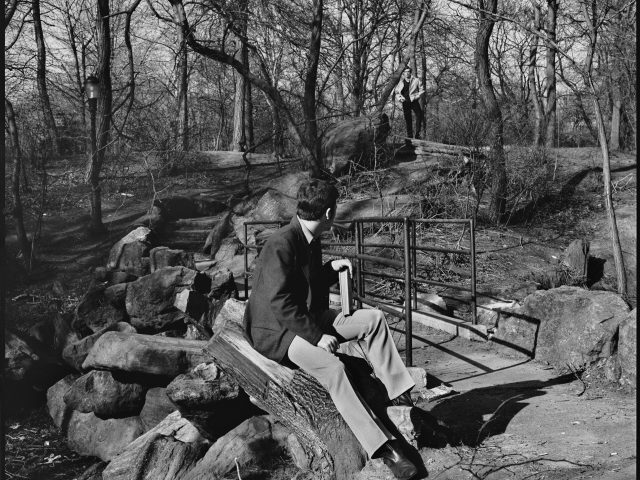 A black and white photograph of a young man sitting on a rock in a park holding a book.