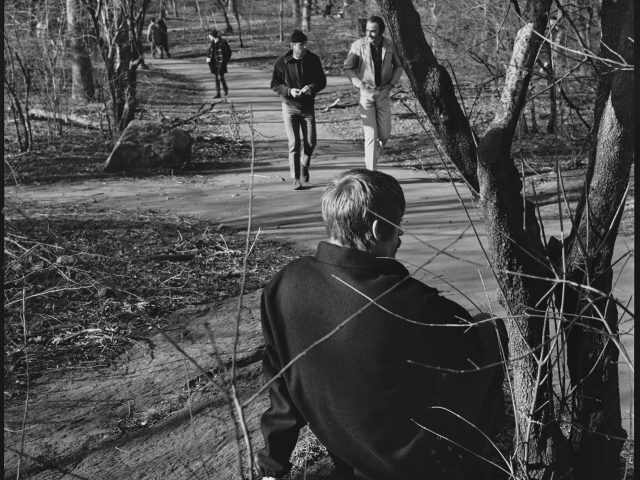 A black and white photograph of a young man sitting in a park in an overcoat.