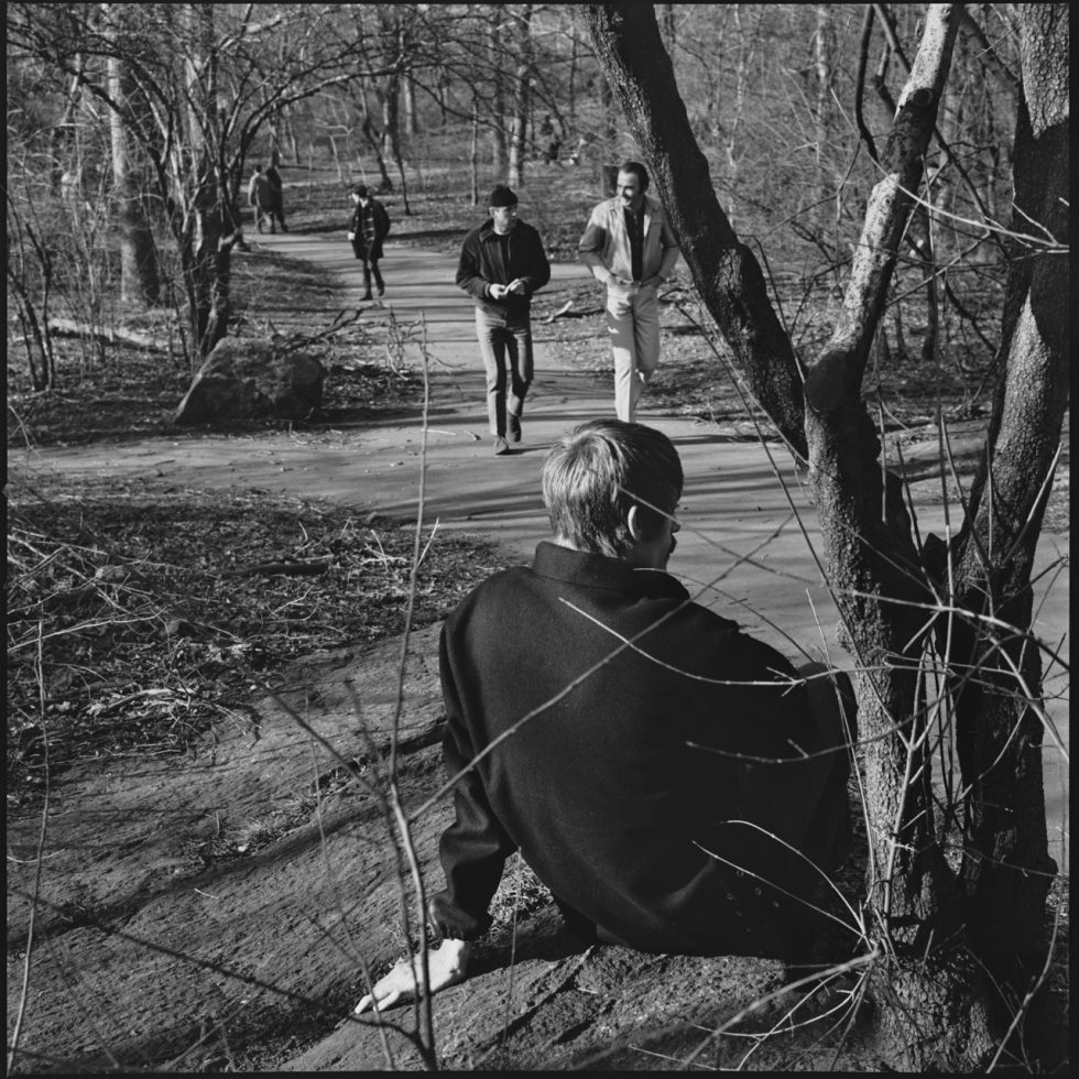 A black and white photograph of a young man sitting in a park in an overcoat.