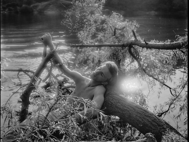 A black and white photograph of a young shirtless man lying on a tree trunk in water.