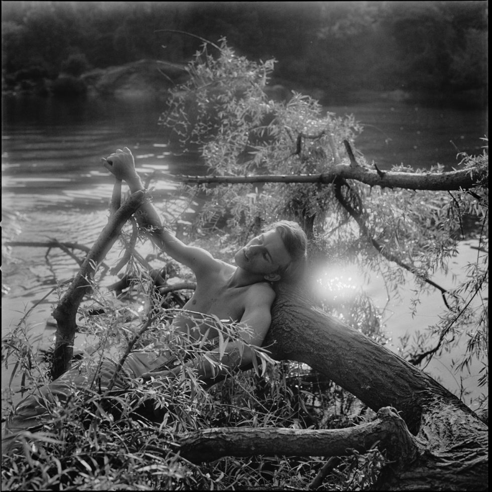 A black and white photograph of a young shirtless man lying on a tree trunk in water.