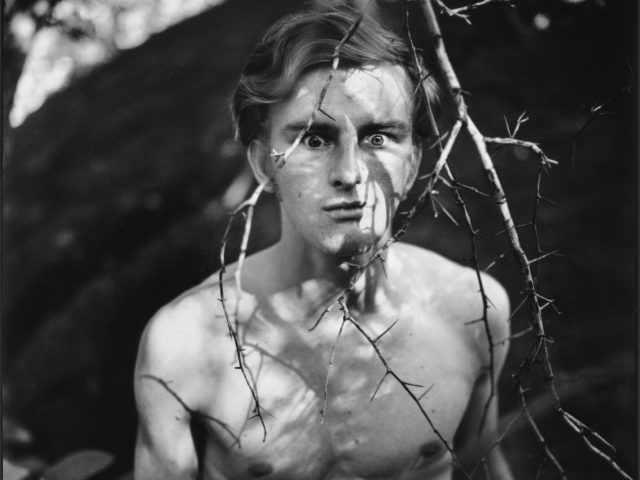 A black and white photograph of a young shirtless man in the woods.