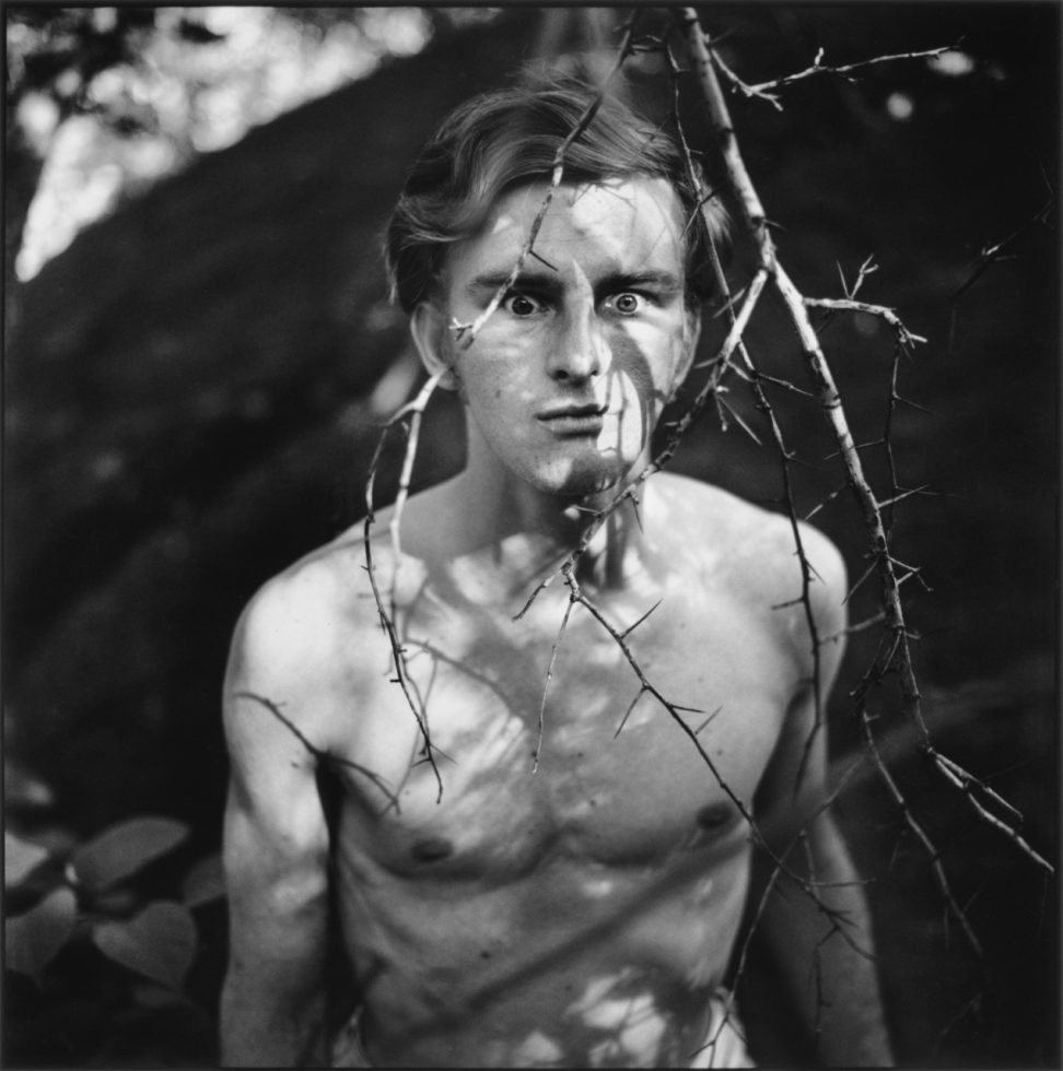 A black and white photograph of a young shirtless man in the woods.